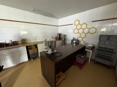 Ein sauberer, moderner Arbeitsbereich mit weiß gefliesten Wänden, auf denen ein Wandbild mit Bienenwaben zu sehen ist, einem dunklen Holztisch, Honiggläsern in Regalen, gestapelten Metallbehältern und Plastikbehältern.