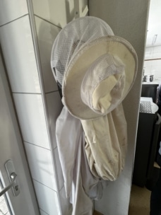 Zwei weiße Imkeranzüge mit Netzhüten hängen an Haken an einer gekachelten Wand in der Nähe einer Tür in einem Raum.