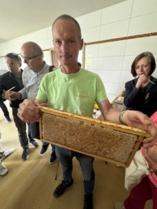 Ein Mann in einem hellgrünen Hemd hält in einem Innenraum einen Wabenrahmen, um den herum mehrere Personen stehen, von denen einige ihn genau beobachten. Im Hintergrund sind weiße Kachelwände zu sehen.