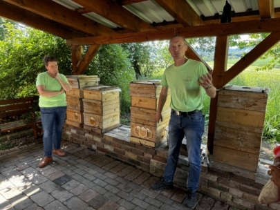 Zwei Personen stehen unter einem hölzernen Unterstand neben gestapelten Holzbienenstöcken. Eine Person spricht mit erhobener Hand, während die andere nachdenklich zuhört. Das Sonnenlicht fällt durch die Bäume im Hintergrund.