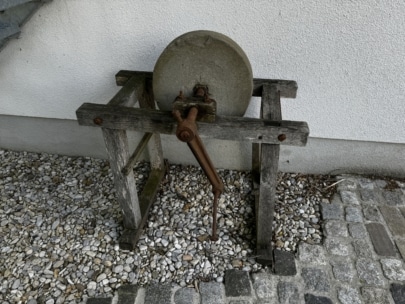 Ein verwitterter, handbetriebener Schleifstein mit einem rostigen Griff in einem Holzrahmen steht auf Kies neben einer weißen Mauer und Steinfliesen.
