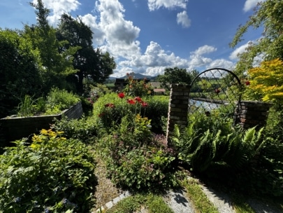 Ein üppiger Garten mit leuchtenden Blumen, grünen Pflanzen und Farnen umgibt einen Steinweg, der zu einem gemauerten Torbogen führt. Im Hintergrund sind ein blauer Himmel mit vereinzelten Wolken und weit entfernte Häuser zu sehen.