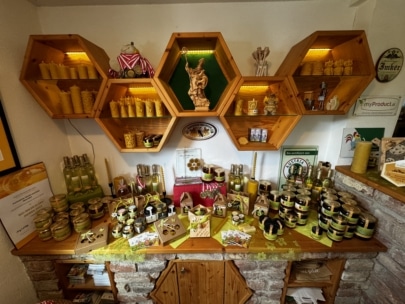 Auf sechseckigen Holzregalen und einem Tisch werden Produkte rund um Honig und Bienen ausgestellt, darunter Gläser, Kerzen, Flaschen und Dekorationsartikel, die in einem gemütlichen Ladenlokal ordentlich angeordnet sind.