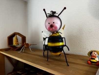 Eine skurrile, bienenförmige Figur mit rundem, rosafarbenem Gesicht und Fühlern steht auf einem Holzregal. In der Nähe befinden sich ein Bienenwaben-Ornament, ein kleines Metallinsekt und eine Puppe mit einem Hut.
