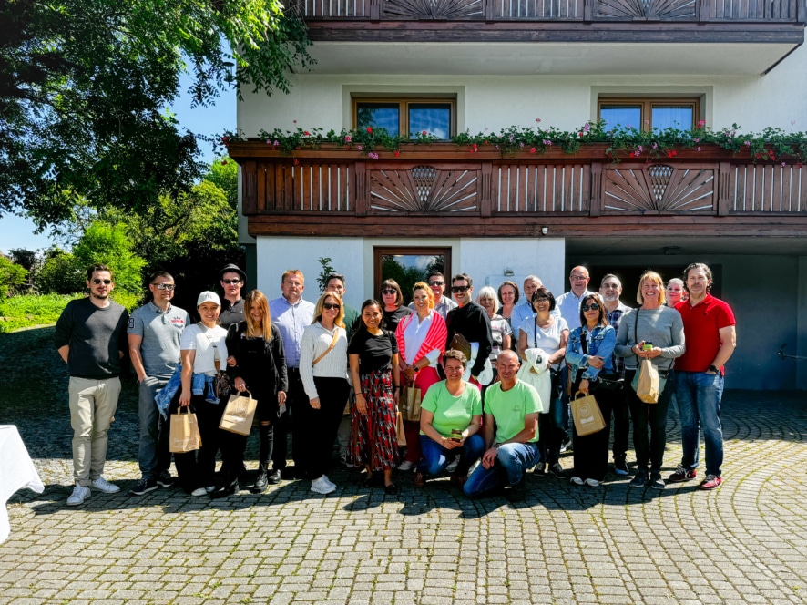 Eine Gruppe von Menschen posiert und lächelt an einem sonnigen Tag im Freien vor einem weißen Gebäude mit Holzbalkonen und Blumenkästen zusammen. Einige halten Papiertüten mit Geschenken; seitlich sind Bäume und Grünpflanzen zu sehen.