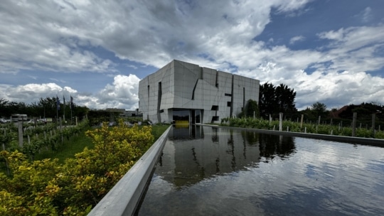 Ein modernes, geometrisches weißes Gebäude mit kantigen Linien steht hinter einem spiegelnden Teich, umgeben von Grün, unter einem teilweise bewölkten Himmel.
