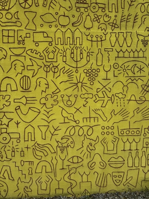Eine leuchtend gelbe Wand, bedeckt mit verschiedenen schwarzen Strichzeichnungen, darunter abstrakte Formen, menschliche Figuren, Tiere, Musikinstrumente, Fahrzeuge, Blumen und Haushaltsgegenstände, die in einem dichten, skurrilen Muster angeordnet sind.
