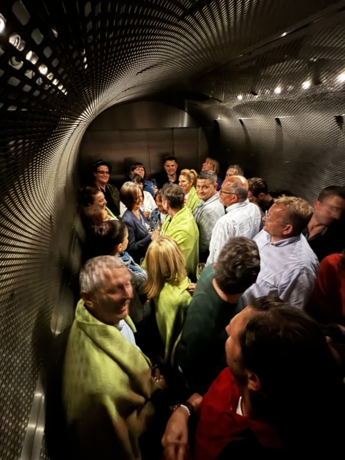 Eine Gruppe von Menschen steht dicht beieinander in einem metallischen, tunnelartigen Raum mit gebogenen Wänden. Einige tragen grüne Schals. Die Atmosphäre wirkt lebendig und gesellig, die Menschen reden und lächeln.