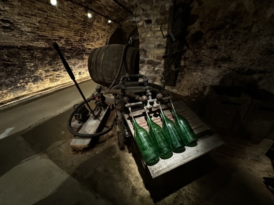 Ein schwach beleuchteter Weinkeller mit einem großen Holzfass auf einer Metallmaschine, vier leeren grünen Glasflaschen, die darunter aufgereiht sind, und Steinmauern im Hintergrund.