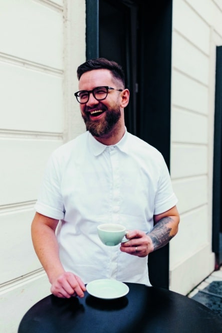 Ein bärtiger Mann mit Brille und weißem Hemd steht draußen an einem schwarzen Tisch, lächelt und hält eine weiße Kaffeetasse. Auf dem Tisch vor ihm steht eine leere Untertasse.