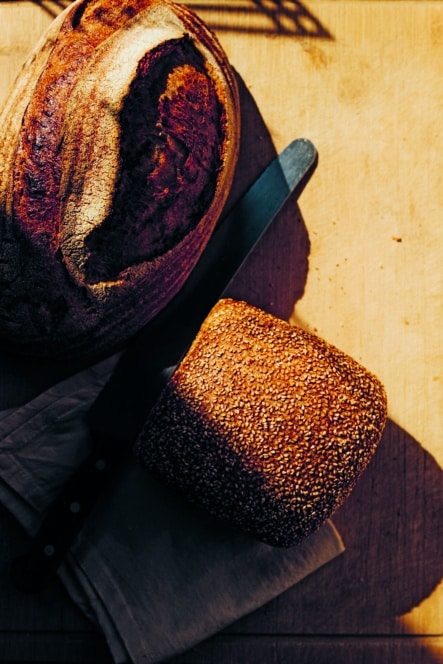 Ein runder Laib Bauernbrot und ein rechteckiger Laib mit Sesamkruste liegen neben einem Brotmesser und einer gefalteten Serviette im warmen Sonnenlicht auf einer Holzplatte.