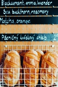 Fünf rustikale Brotlaibe in einem weißen Drahtkorb liegen unter einer Menütafel, auf der Geschmacksrichtungen wie Schwarze Johannisbeere, Aronia, Lavendel, Sanddorn, Rosmarin, Matcha und Orange aufgeführt sind, darunter ein tschechisches Brotetikett.