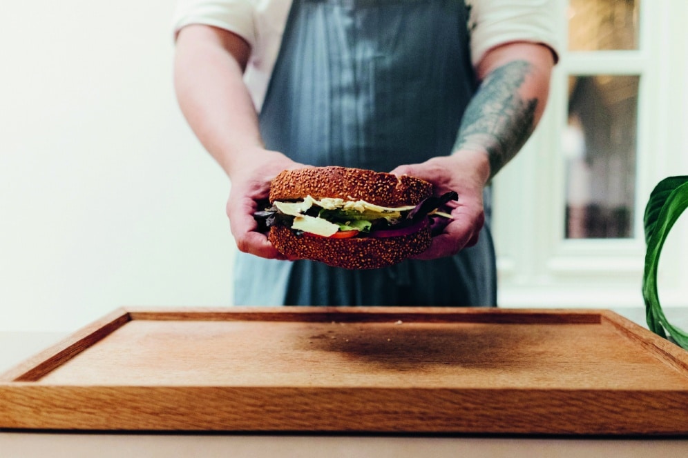 Eine Person in einer Schürze hält ein großes Sandwich mit Salat, Käse und Tomate über ein hölzernes Schneidebrett, wobei sie die Arme in Richtung Kamera ausstreckt.