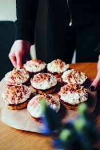 Eine schwarz gekleidete Person hält ein Holzbrett mit acht Cupcakes auf einem Holztisch, die mit weißem Zuckerguss und zerbröseltem Belag verziert sind.