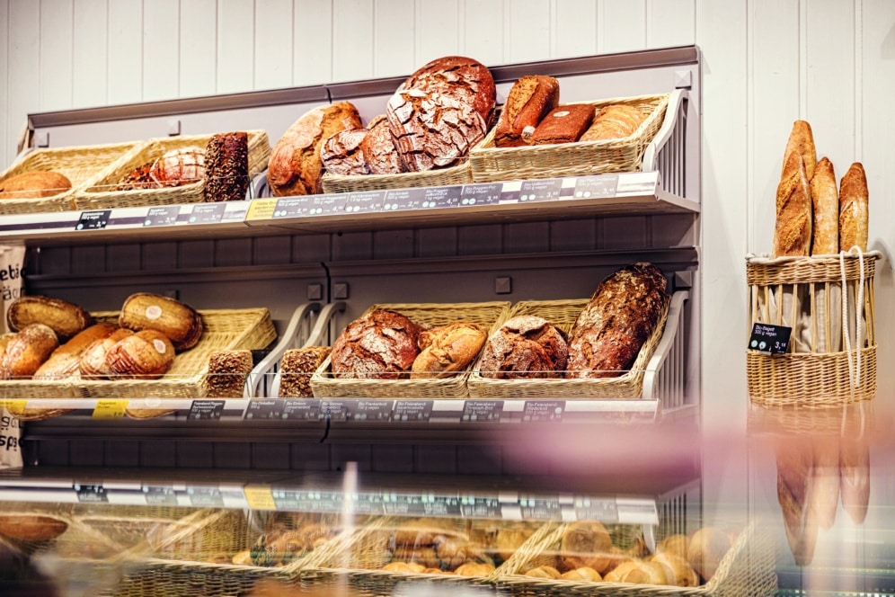 In einer Bäckerei stehen mehrere Körbe mit verschiedenen Arten von frischem Brot und Baguette in Regalen, im Vordergrund sind eine Theke und weitere Backwaren zu sehen.