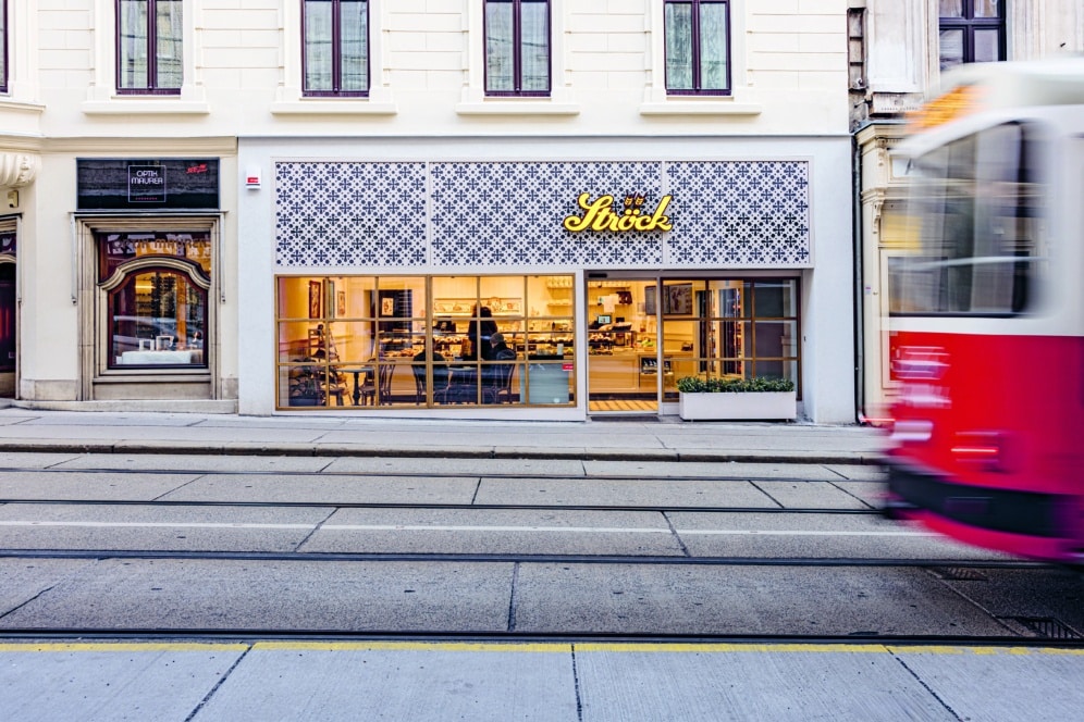Straßenansicht eines modernen Cafés mit gemusterten Kacheln über großen Fenstern, warmer Beleuchtung im Inneren und einer unscharfen rot-weißen Straßenbahn, die auf Gleisen im Vordergrund vorbeifährt.