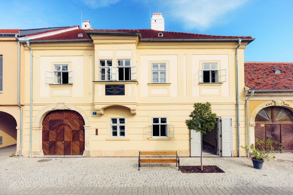 Ein cremefarbenes historisches Gebäude mit Holztüren, fünf offenen Fenstern, einer Bank und einem Topfbaum auf einer Kopfsteinpflasterstraße unter einem klaren blauen Himmel.