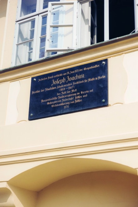 Eine Joseph Joachim gewidmete Gedenktafel ist an einer hellgelben Hauswand unter einem offenen Fenster angebracht. Die Tafel enthält einen Text in deutscher Sprache über seine Leistungen und seine Geburt.