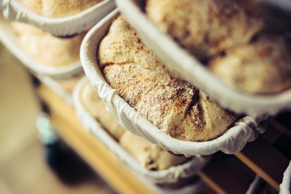 Nahaufnahme mehrerer Teiglaibe, die in mit Mehl bestäubten Gärkörben aufgehen und sich wahrscheinlich darauf vorbereiten, zu Brot gebacken zu werden. Die Körbe sind auf Holzgestellen gestapelt.
