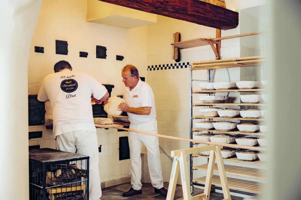 Zwei Bäcker in weißen Uniformen arbeiten in einer rustikalen Bäckerei und bereiten Brotlaibe vor. Einer schiebt das Brot in den Ofen, während der andere mit dem Teig hantiert. An der Wand neben ihnen stehen Regale mit Brottabletts.