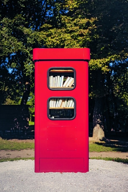 Ein leuchtend roter Büchertauschkasten im Freien mit zwei Fenstern, in denen Bücher ausgestellt sind, steht auf einem Kiesplatz mit Bäumen und Grün im Hintergrund.
