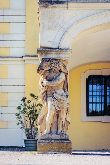 Eine steinerne Statue einer gewandeten Figur ist in eine Gebäudesäule integriert und stützt einen Torbogen. Die Fassade des Gebäudes ist hellgelb mit weißer Umrandung, und in der Nähe befindet sich eine Topfpflanze mit rosa Blüten.