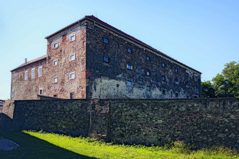 Ein großes, altes Backsteingebäude mit vielen kleinen Fenstern steht an einem sonnigen Tag hinter einer hohen Steinmauer. Das Gebäude weist Anzeichen von Verwitterung und Alter auf, die Fassade ist stellenweise verblasst und blättert ab.