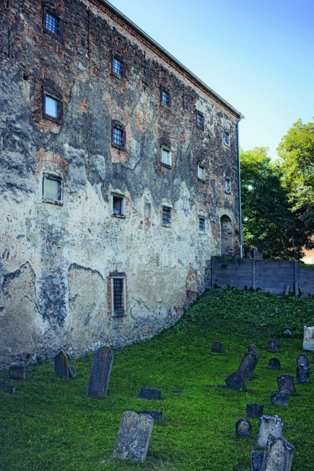 Ein altes Steingebäude mit verwitterten Mauern steht neben einem grasbewachsenen Friedhof mit mehreren schiefen und abgenutzten Grabsteinen; im Hintergrund stehen Bäume und ein Holzzaun unter einem klaren blauen Himmel.