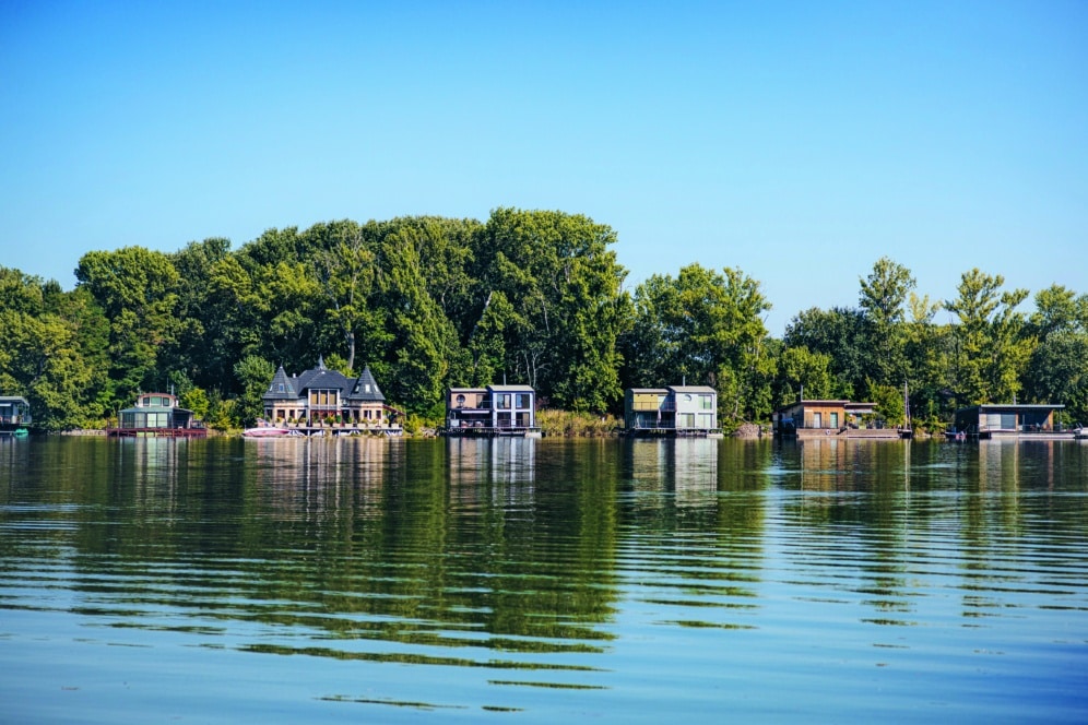 Eine Reihe von Häusern und Hausbooten liegt an einem grünen, von Bäumen gesäumten Ufer, das sich im ruhigen Wasser eines Sees unter einem klaren blauen Himmel spiegelt.