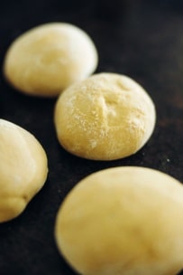 Vier runde, goldene Brötchen liegen auf einer dunklen Fläche. Die Brötchen sind leicht mit Mehl bestäubt und haben auf der Oberseite eine leicht rissige Textur, was darauf hindeutet, dass sie frisch gebacken sind.