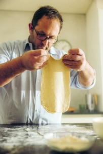 Ein Mann mit Brille streckt den Pizzateig in seinen Händen über einer bemehlten Arbeitsfläche in einer sonnenbeschienenen Küche aus, während im Hintergrund eine Uhr und Utensilien zu sehen sind.