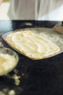 Ein rechteckiger Pizzateig, bestrichen mit einer Schicht cremiger Ricotta, liegt auf einer dunklen Arbeitsplatte, daneben eine Schüssel mit noch mehr Ricotta.