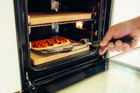 Eine Hand benutzt einen Pizzaschieber, um eine rohe Pizza auf einen heißen Pizzastein in einem offenen Ofen zu legen. Die Pizza ist mit Tomaten und Oliven belegt. Das Licht des Ofens ist eingeschaltet und beleuchtet die Szene.