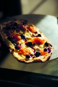 Ein rechteckiges Fladenbrot mit leuchtend orangefarbenen, gelben und violetten Zutaten, möglicherweise Obst und Gemüse, liegt auf einem Backblech, das von warmem Sonnenlicht angestrahlt wird.