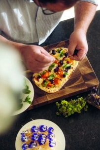 Eine Person garniert ein Fladenbrot mit frischen Kräutern, Tomaten und essbaren Blüten auf einem Holzbrett. In der Nähe steht ein Teller mit Spießen, die mit blauen und weißen essbaren Blumen belegt sind, und auf der Theke stehen frische grüne Kräuter.