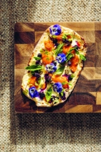 Ein rechteckiges Fladenbrot mit geräuchertem Lachs, Frischkäse, Kräutern, Gemüse und blauen essbaren Blüten liegt auf einem hölzernen Schneidebrett, das mit einer strukturierten Oberfläche versehen ist.