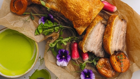 Schweinebraten mit knuspriger Haut, in Scheiben geschnitten, um das saftige Fleisch freizulegen, serviert mit Aprikosen, Radieschen, essbaren lila Blüten, Grünzeug und einer Schale mit grüner Soße auf Pergamentpapier.