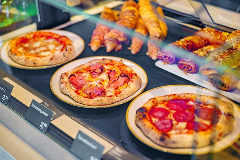 Drei Pizzateller mit verschiedenen Belägen, darunter Peperoni und Schinken, werden in einer gläsernen Bäckereivitrine neben verschiedenen Arten von Backwaren im Hintergrund ausgestellt. Die Preisschilder sind vor den Pizzen sichtbar.