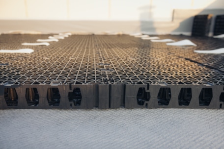 Nahaufnahme von schwarzen Kunststoff-Drainageplatten, die auf einer ebenen Fläche verlegt wurden, möglicherweise für ein Gründach oder ein Bodensystem, mit geometrischen Mustern und weißem Material, das teilweise darunter sichtbar ist.