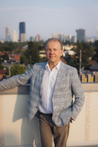 Ein Mann in einem hellgrauen Blazer und einem weißen Hemd steht an einem sonnigen Tag auf einem Dach mit einer Stadtsilhouette und hohen Gebäuden im Hintergrund.