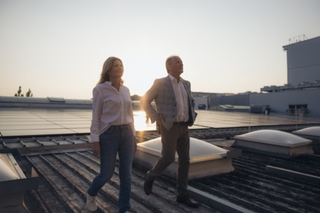 Zwei Personen, eine Frau und ein Mann, gehen bei Sonnenuntergang auf einem Dach mit Sonnenkollektoren spazieren. Der Himmel ist klar, und das Sonnenlicht scheint hinter ihnen. Die Atmosphäre lässt auf eine ruhige, professionelle Umgebung schließen.