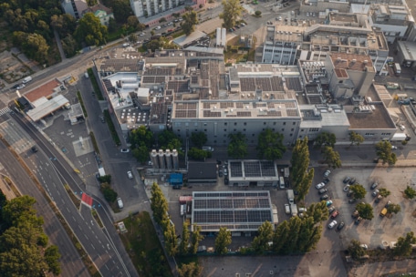 Luftaufnahme eines Industriekomplexes mit mehreren Gebäuden und vielen mit Solarzellen bedeckten Dächern, umgeben von Bäumen, Straßen, parkenden Autos und der angrenzenden städtischen Infrastruktur.