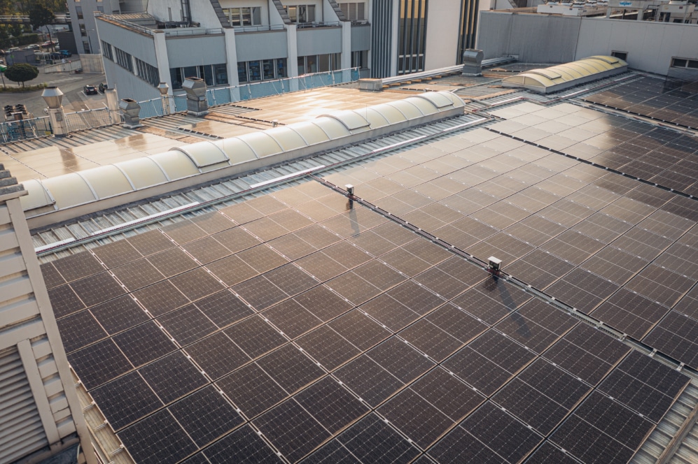Eine große Anordnung von Sonnenkollektoren auf dem Flachdach eines Industriegebäudes, die das Sonnenlicht reflektieren. In der Nähe befindliche Strukturen und Lüftungsanlagen sind ebenfalls zu sehen.