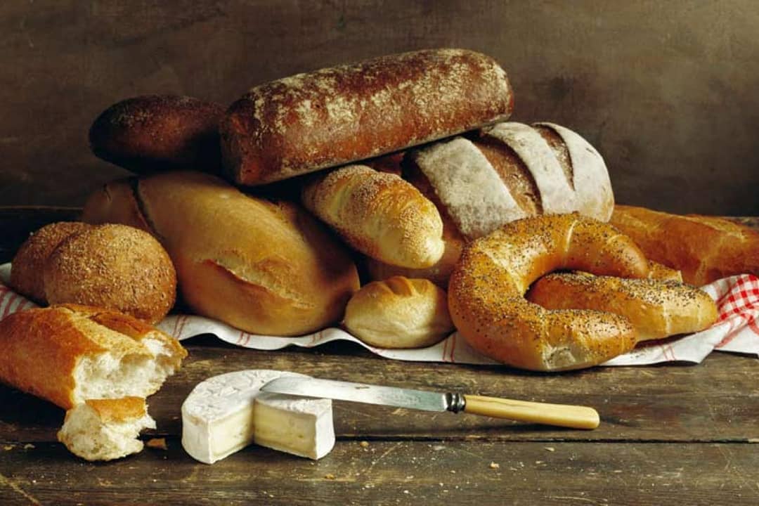 Eine Auswahl an Broten, darunter Brötchen, Baguettes, ein Mohnbagel und geschnittene Brote, sind auf einem Tuch angeordnet. Davor liegen ein Stück Weichkäse und ein Messer auf einem rustikalen Holztisch.