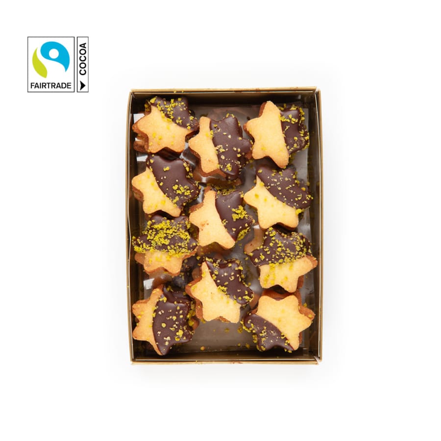 Eine Schachtel mit sternförmigen Keksen, halb mit Schokolade überzogen und mit gehackten Pistazien bestreut. In der oberen linken Ecke befindet sich ein Etikett mit Fairtrade-Kakao. Die Kekse sind ordentlich auf einem weißen Hintergrund angeordnet.