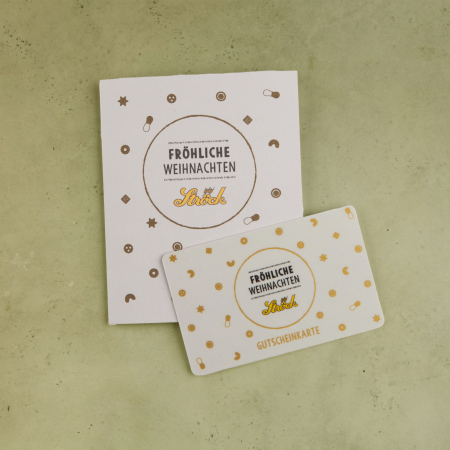 Eine weiße Geschenkkarte und ein passender Umschlag mit goldenen Festtagssymbolen und dem Text Fröhliche Weihnachten Stöck GUTSCHEINKARTE auf beiden, platziert auf einer grün strukturierten Oberfläche.