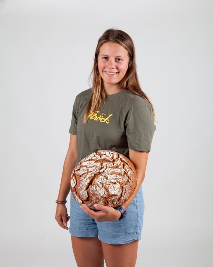 Eine junge Frau mit langen braunen Haaren, die ein grünes T-Shirt und blaue Shorts trägt, lächelt, während sie einen runden, mit Mehl bestäubten Laib rustikalen Brotes vor einem schlichten weißen Hintergrund hält.