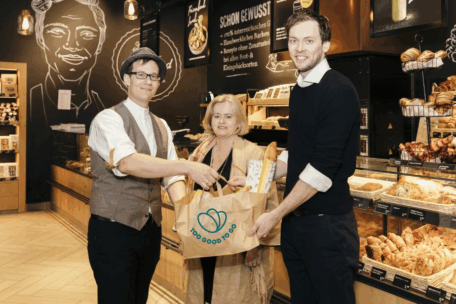 Drei Personen stehen in einer Bäckerei und überreichen eine Papiertüte mit der Aufschrift "Too Good To Go", die mit Brot und Lebensmitteln gefüllt ist. Im Hintergrund sind Gebäck und Kreidetafeln zu sehen.