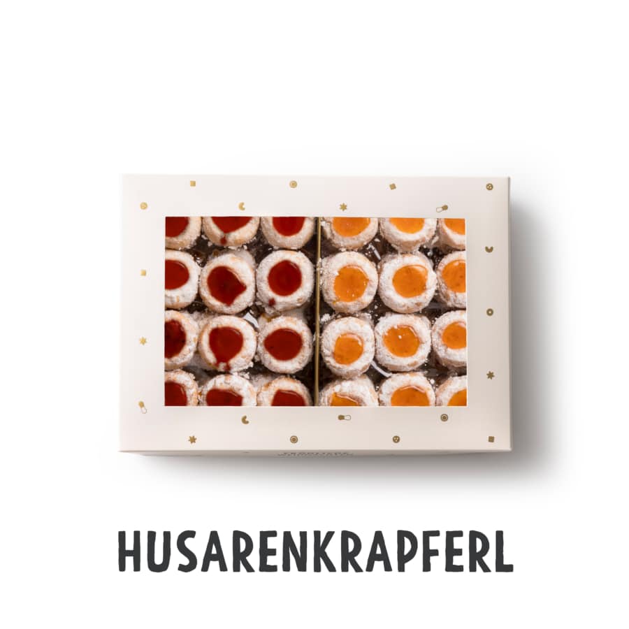 Eine Schachtel mit Husarenkrapferl, runde Kekse, gefüllt mit roter und oranger Marmelade, in ordentlichen Reihen angeordnet. Die Schachtel ist weiß mit goldenen Sternen und Monden verziert, darunter steht HUSARENKRAPFERL.