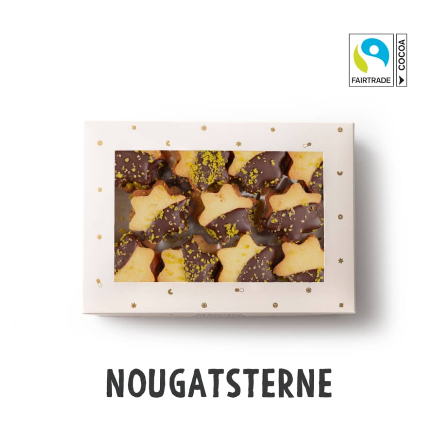 Abgebildet ist eine Schachtel mit sternförmigen Keksen, die halb in Schokolade getaucht und mit zerstoßenen Pistazien bestreut sind. Die Verpackung hat ein durchsichtiges Fenster und das Fairtrade-Kakao-Logo ist in der oberen rechten Ecke zu sehen. Darunter steht NOUGATSTERNE geschrieben.